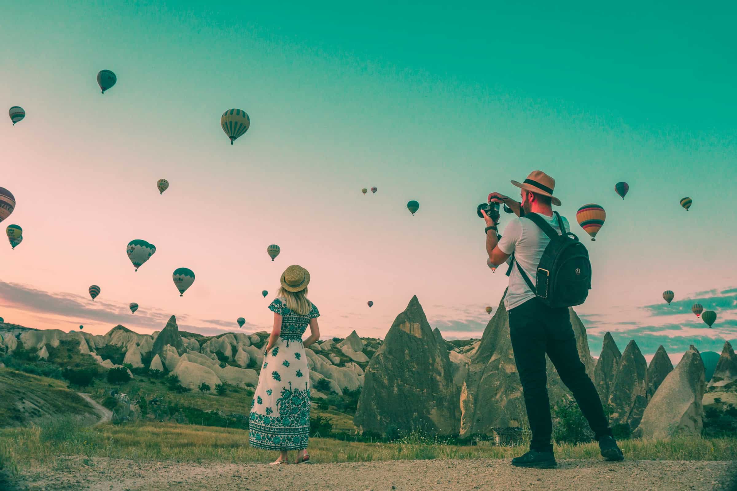 anniversaries-img-02 Balloons floating over unique rock formations in Cappadocia during sunrise with a photographer capturing the scene, illustrating dream travel destinations and adventure photography.