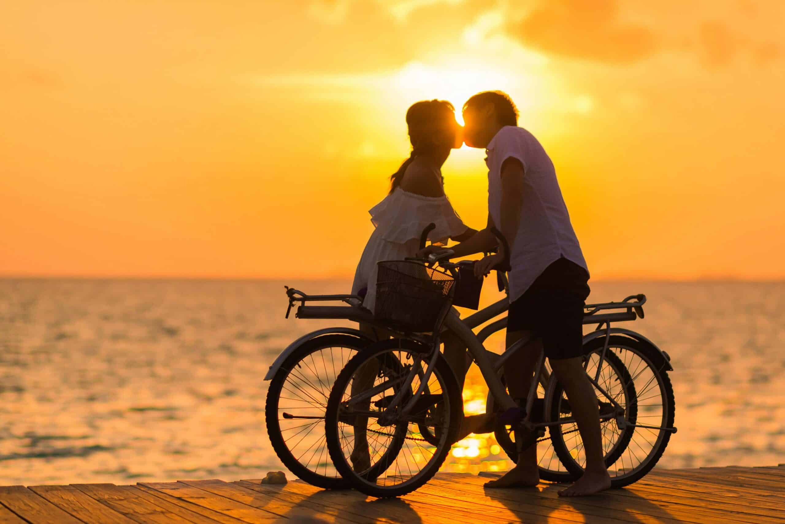 Silhouettes of a couple kissing at sunset on a beach with bicycles, romantic vacation, summer travel, beach getaway, couple's trip, destination wedding, tropical beach, sunset romance, travel adventure, travel agency.