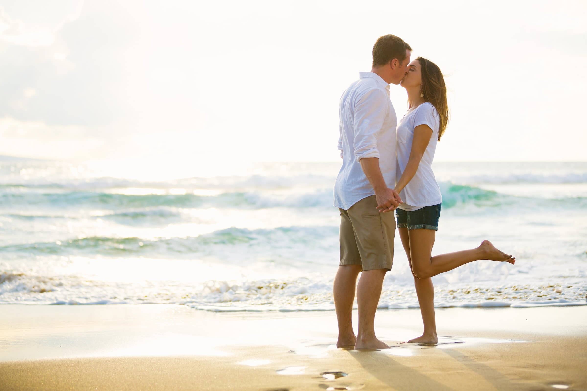 Romantic couple kissing on the beach at sunset, wearing casual white shirts and shorts, celebrating love and destination weddings, perfect for honeymoon or vacation travel photography.