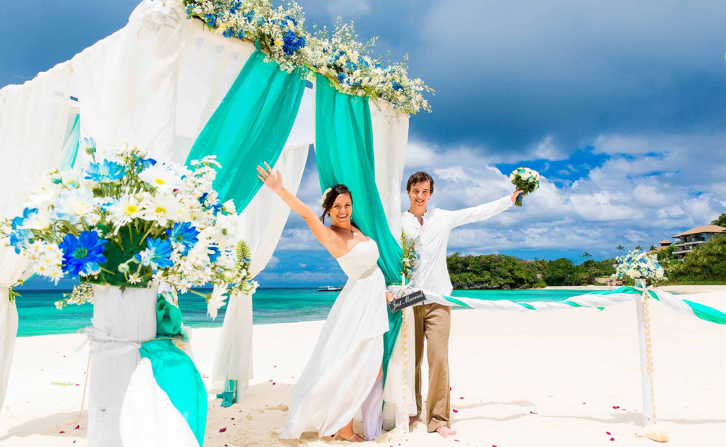 Vibrant beach wedding ceremony with a happy bride and groom celebrating under a turquoise and white floral arch on a sunny tropical beach.