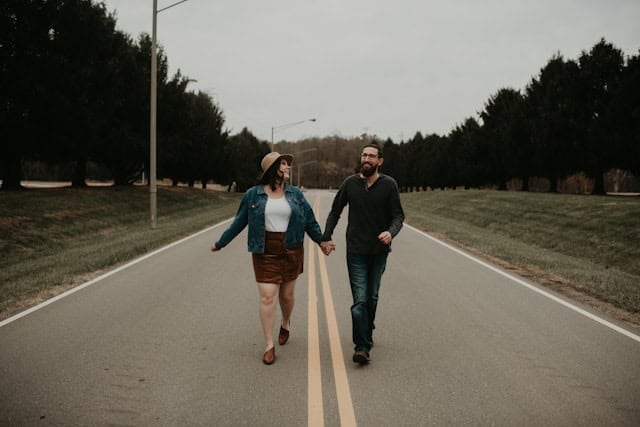 A couple walking hand in hand down a quiet rural road, celebrating their engagement and love, with lush trees on either side, under overcast skies.