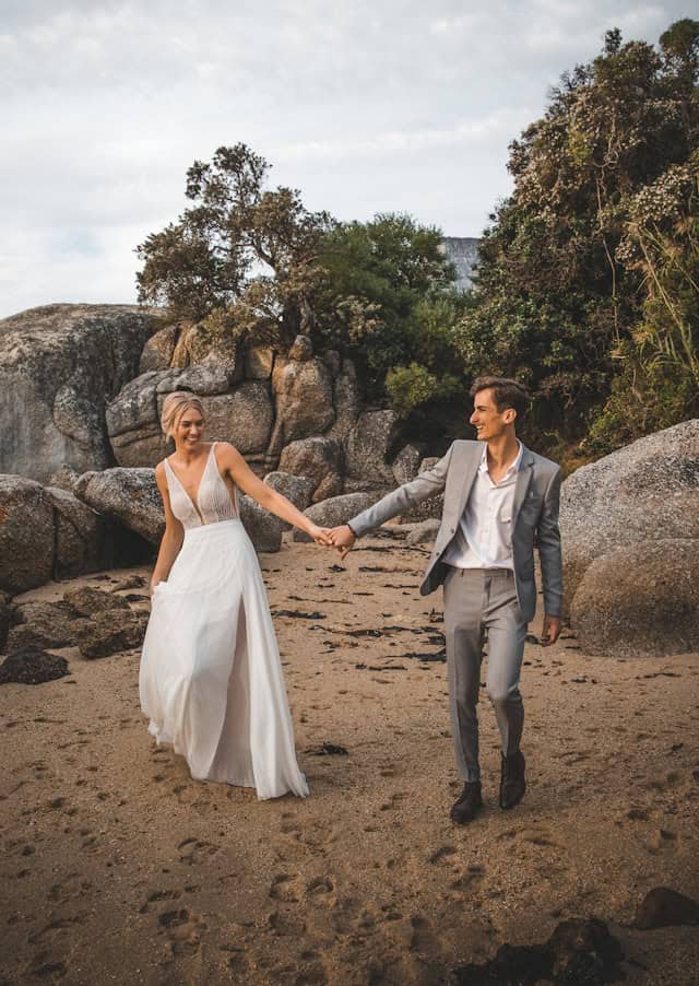 Romantic couple in wedding attire walking on the beach at a scenic destination, celebrating love and destination weddings, with natural rocky landscape and trees in the background.
