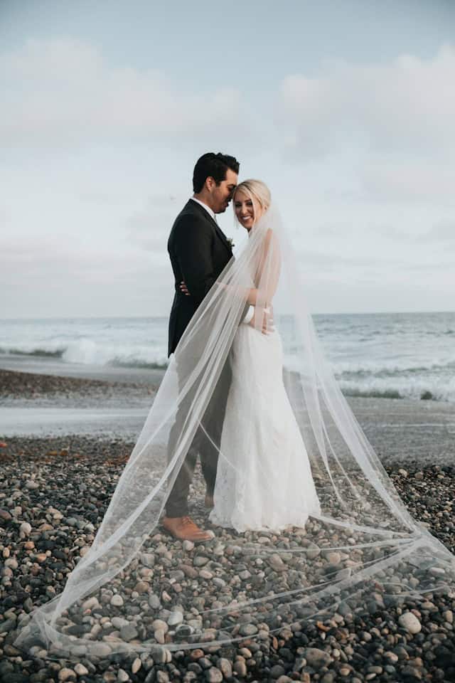 Romantic beach wedding couple in wedding dress and tuxedo under cloudy sky, bride with veil and groom smiling, ocean backdrop, wedding photography by I Do Destinations by vivid, destination wedding planning, beach wedding photos.