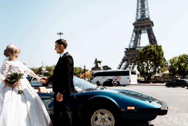 anniversaries-img-02 Couple in wedding attire next to a classic sports car with the Eiffel Tower in the background in Paris, France, representing romantic destination weddings and luxury travel.