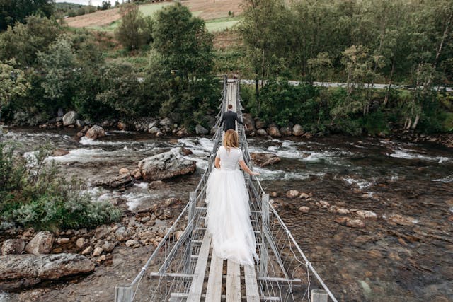 anniversaries-img-02 Elegant bride walking across a rustic suspension bridge over a mountain river, capturing adventurous wedding destination moments, outdoor wedding venue ideas, and scenic wedding photography.