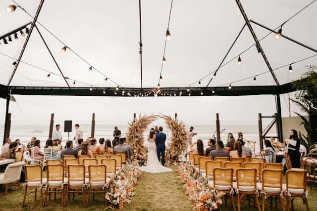 anniversaries-img-02 A1. Beachside wedding ceremony setup with floral arch and ocean view, outdoor wedding venue with guests seated under string lights, romantic destination wedding by I Do Destinations by vivid.