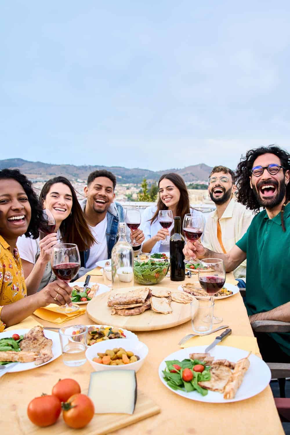 Group of friends enjoying a meal outdoors with wine and food, celebrating together with smiles and laughter.