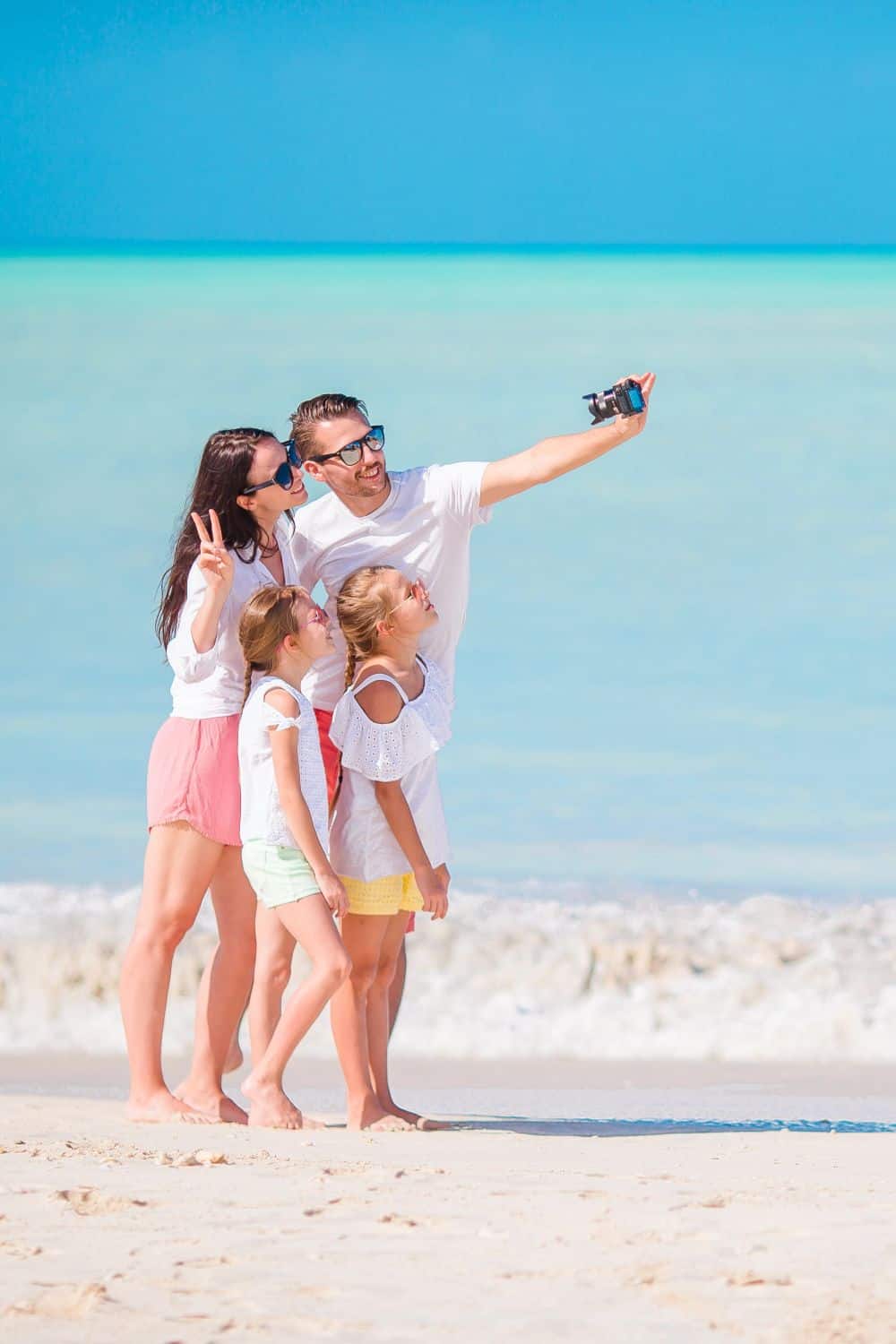 Family of four taking a selfie on a sunny beach with clear blue water and sky in the background, enjoying a vacation together.