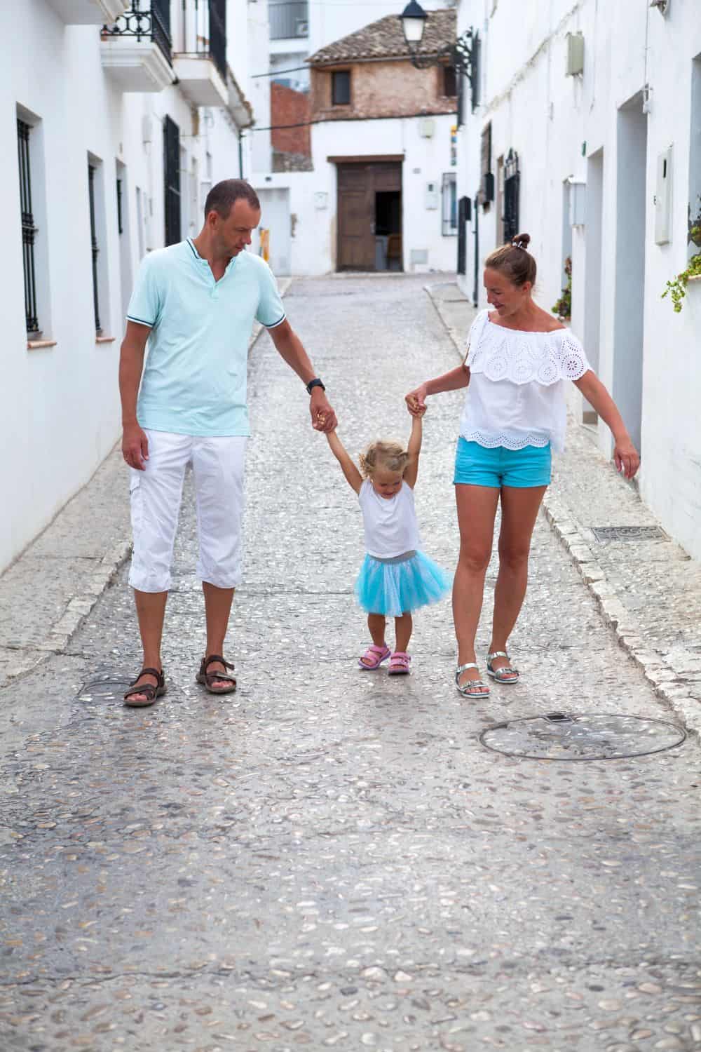 Family walking hand-in-hand through a charming, cobblestone street in a historic town with white buildings and narrow alleyways.