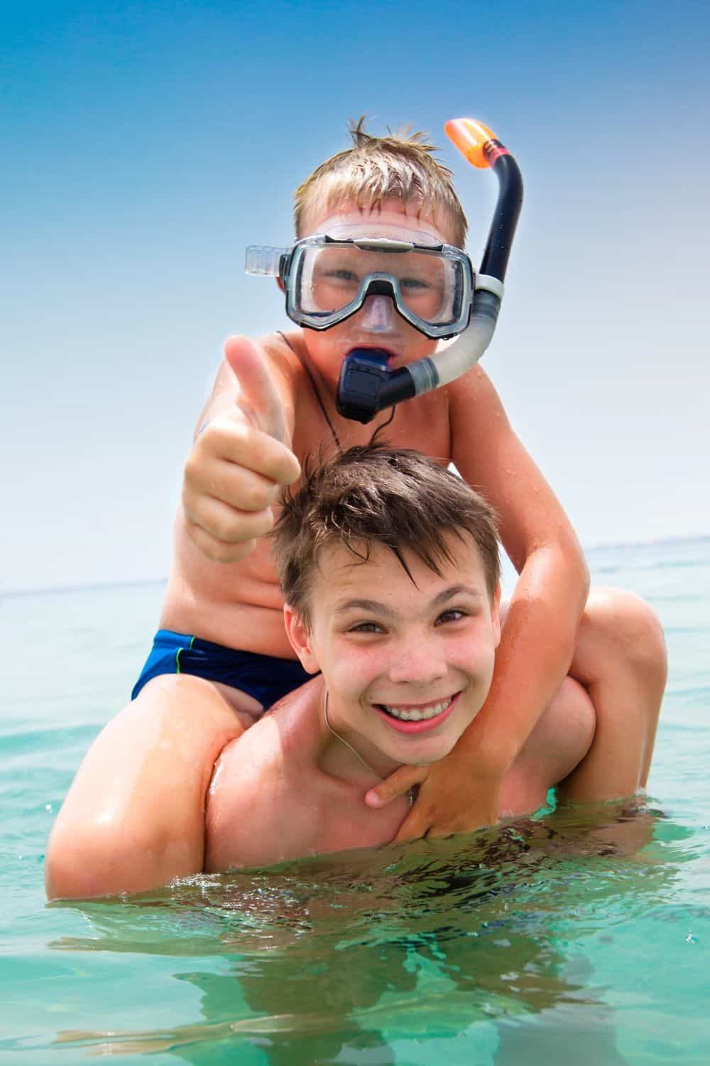 Two children enjoying snorkeling in clear ocean water, one wearing a snorkel mask and giving a thumbs-up, capturing a fun family beach activity.
