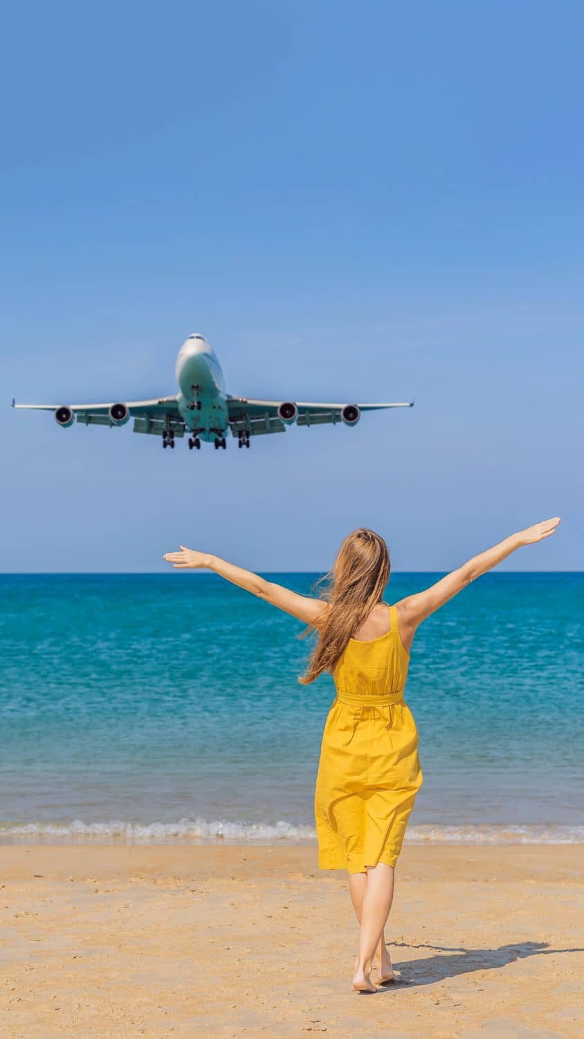 Woman in yellow dress on beach with arms outstretched, airplane flying overhead in clear blue sky, ocean in background.